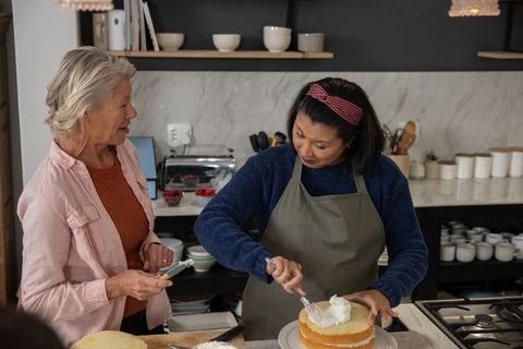 Older Caucasian and Asian Friends Decorating Layered Cake in Cozy Home Kitchen