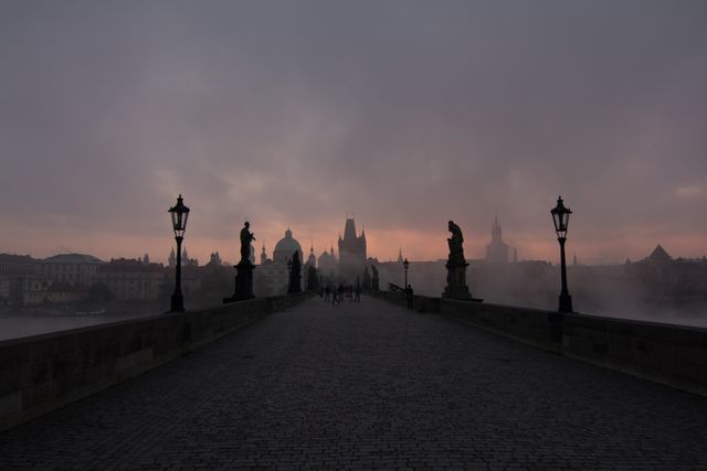 Scenic view of an old European stone bridge during misty dawn with silhouetted statues and lampposts lining the cobblestone path. The fog and sunrise create an atmospheric and tranquil scene, highlighting historical architecture. Ideal for travel blogs, promotional materials for Europe travel, and decorative wall art.