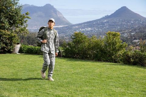 Asian soldier walking on grassy hillside carrying tactical backpack with mountain valley