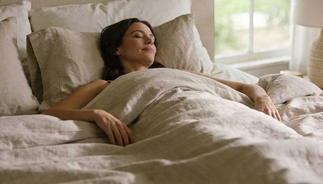 Woman resting in sunlit bed relaxing under neutral linen duvet minimalist bedroom calm
