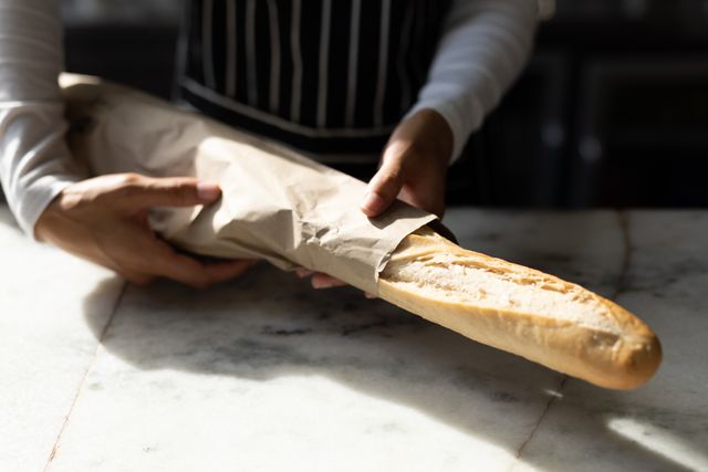 Midsection of a biracial young female barista holding a freshly baked baguette wrapped in paper at a cafe counter. Ideal for use in articles or advertisements related to cafes, bakeries, food service, and culinary arts. Can also be used to illustrate concepts of fresh food, customer service, and artisanal baking.