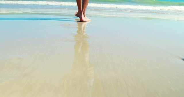 Barefoot person casually walking by the seashore on a sunny day. The feet and lower legs are reflected in the wet sand, with gentle waves in the background. Ideal for themes such as relaxation, summer vacation, seaside leisure, serenity, and tranquil coastal scenes.