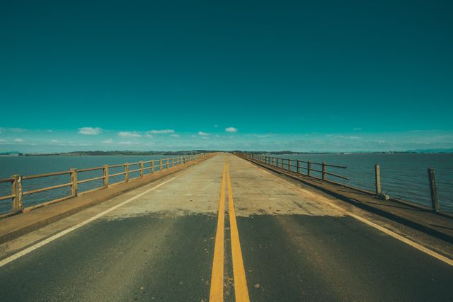 Symmetrical deserted road on an aged asphalt bridge extending over calm waters. Majestic horizon view with clear sky creating a serene and tranquil atmosphere. Ideal for illustrations of solitude, journey, emptiness, or calmness. Useful for travel blogs, inspirational posters, road trip advertisements, and more.