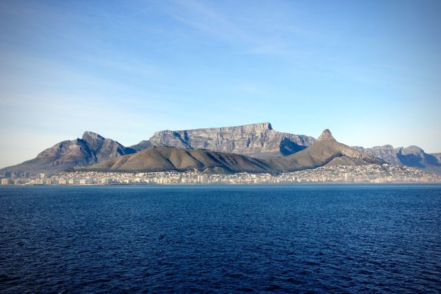 Features the renowned Table Mountain with its distinctive flat top. In the foreground, the calm blue ocean contrasts with the rugged mountain landscape. The sky is clear, enhancing the natural beauty and expanse of this South African landmark. Ideal for travel blogs, tourism promotions, educational material about geography, or desktop backgrounds.