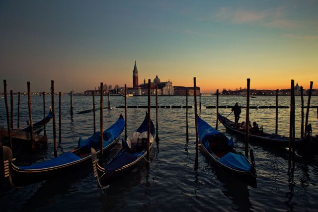 Gondolas resting on a canal in Venice at sunset with a cityscape and a serene horizon. Picture perfect for romantic travel advertisements, websites promoting Italy, postcards, and blogs focused on travel and leisure.