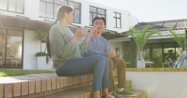 Two friends enjoying a sunny day, sitting on steps, engaging in casual conversation outside a modern home. Ideal for use in lifestyle blogs, real estate websites, social media postings about friendship, and advertisements highlighting a relaxed and joyful atmosphere.