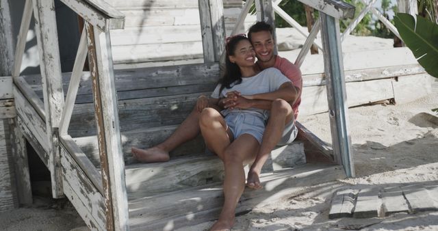Happy couple sitting on rustic wooden steps at the beach, smiling and enjoying each other's company. Suitable for themes of love, romance, summer vacation, relaxation, lifestyle, and travel promotions.