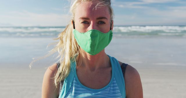 Woman standing on beach wearing a green facemask for outdoor safety. Scene includes sunny day with blue sky and gentle waves in background. Used for promoting health measures, exercise during pandemic, COVID-19 prevention, or stories about outdoor activity with social distancing.