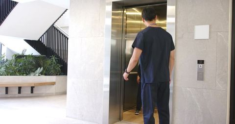 Asian healthcare worker entering elevator in hospital lobby wearing scrubs and id badge