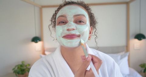 African american woman applying pale green facial mask in cozy bedroom robe for self care