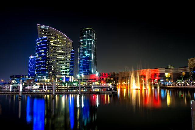 City skyline at night with brightly illuminated modern buildings reflecting in a water body. The vibrant lighting includes various colors. Perfect for concepts related to urban life, modern architecture, travel destinations, and nightlife.