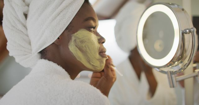 Woman with facial mask in bathroom looking at herself in lighted vanity mirror, focusing on skincare and relaxation. Perfect for content about self-care, beauty routines, spa treatments, and wellness concepts.