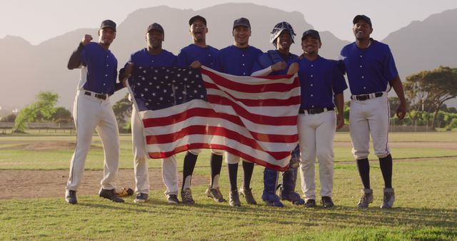 A diverse group of baseball players in blue uniforms celebrating their victory by proudly holding an American flag on a baseball field with mountains in the background. Ideal for use in sports-related advertising, promotion of team spirit and inclusion, or marketing campaigns focusing on American culture and unity.