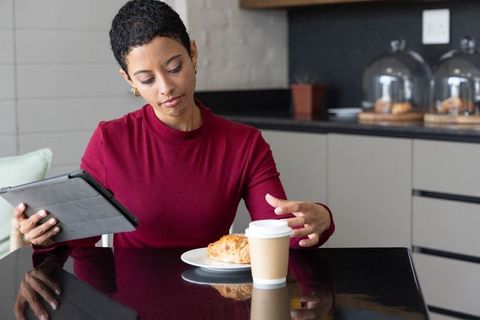 African american woman working on tablet in cafe reaching for takeaway coffee and croissant