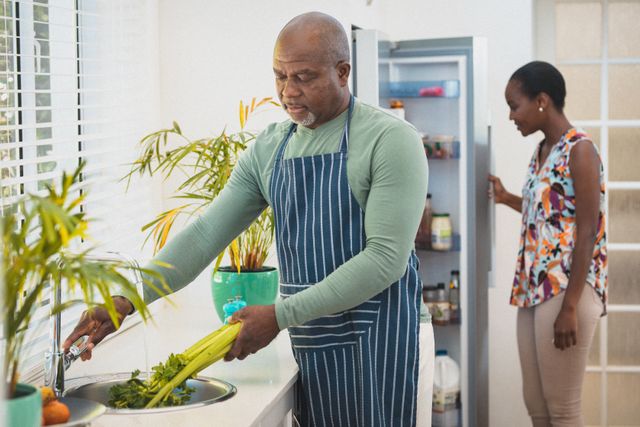 African american senior couple in kitchen male washing vegetables female looking into fridge. at home in isolation during quarantine lockdown.