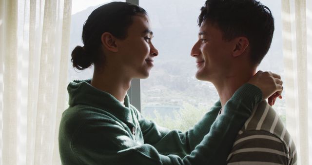 A romantic couple embraces near a window with sunlight filtering through behind them, creating a serene and tender atmosphere. Both individuals are smiling, exhibiting a loving and affectionate connection. Ideal for use in articles or campaigns promoting love, relationships, intimacy, or LGBTQ+ inclusivity.