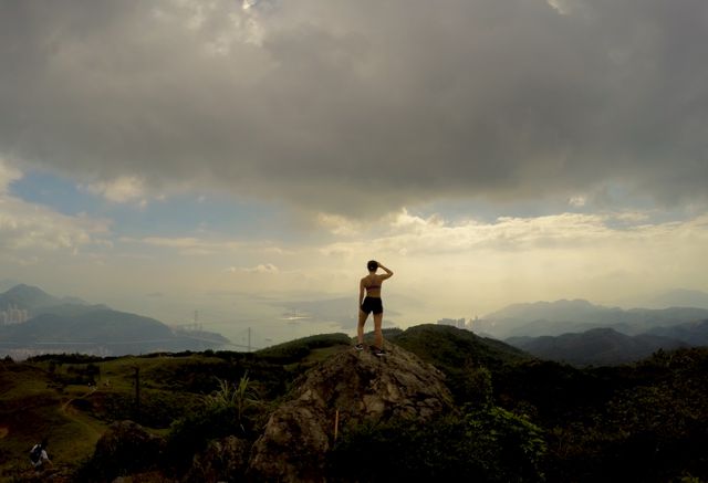 Person standing on rocky mountain peak, overlooking expansive landscape with beautiful sunset. Dramatic sky with clouds enhances the serene and awe-inspiring atmosphere. Perfect for use in travel promotions, adventure advertising, nature documentaries, and motivational visuals focused on exploration and personal achievement.