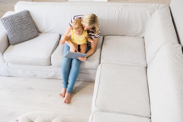 Mother and baby girl sitting on a light gray sofa in a modern living room, using a digital tablet. Ideal for use in articles about parenting, family bonding, technology in daily life, or home lifestyle.