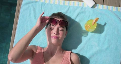 Sunbathing woman lifting sunglasses on pool deck with citrus cocktail and smartphone nearby