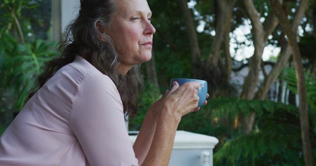 Thoughtful senior caucasian woman standing on terrace drinking coffee and looking around. retirement lifestyle at home alone.