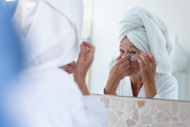 Mature caucasian woman using under eye treatment patch looking in bathroom mirror, copy space. Self care, health, beauty and senior lifestyle concept.