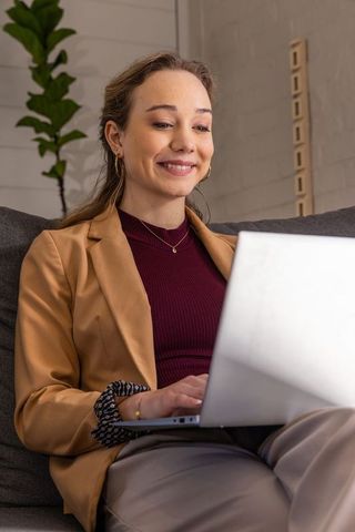 Smiling professional woman working on laptop on couch in business casual attire, modern home office