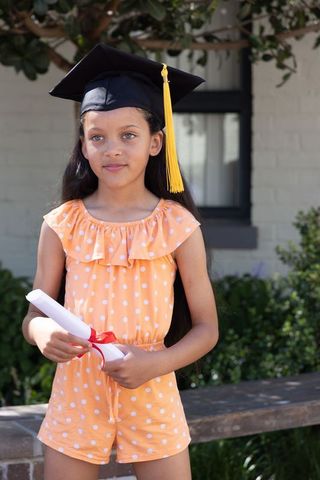 Young girl celebrating graduation wearing mortarboard and holding diploma outdoors