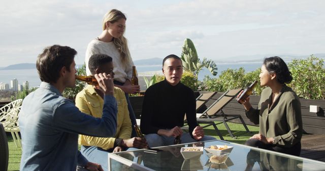 Multiracial group of friends sitting and drinking together outdoors on a sunny day. They are enjoying each other's company while chatting and snacking. Ideal for illustrating social events, friendship, relaxation, and outdoor gatherings.