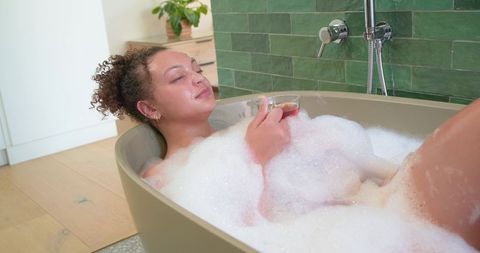 African american woman relaxing in freestanding bathtub with bubbles and glass of tea