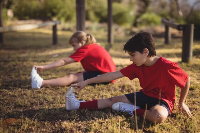 Children in red shirts stretching on grass during an outdoor boot camp obstacle course. Ideal for use in articles or advertisements about children's fitness, outdoor activities, summer camps, physical education, and promoting a healthy lifestyle for kids.