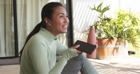 Mid 30s asian woman smiling and relaxing on sunlit porch holding smartphone and pink bottle
