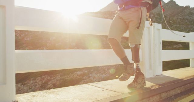 Low section of biracial man with prosthetic leg trekking with backpack crossing bridge, backlit. Long distance walking, fitness, challenge, disability and healthy outdoor lifestyle.