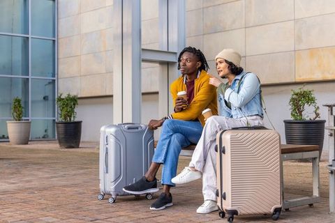 Urban travelers resting on bench with coffee and rolling luggage waiting at plaza
