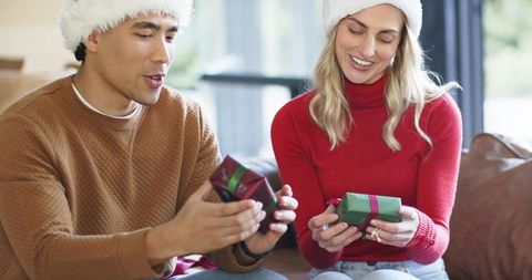 Couple exchanging small wrapped gifts on sofa wearing santa hats and cozy sweaters