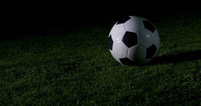A classic black and white soccer ball rests on a lush green field at night, illuminated by a focused light, with copy space. Its position suggests a paused moment in a game or practice session.