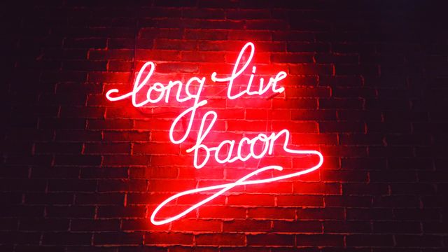 Neon sign saying 'Long Live Bacon' glowing brightly on dark brick wall. Perfect for restaurant websites, diner advertisements, kitchen decor inspirations, and retro aesthetic marketing materials.