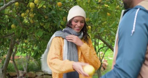 Couple sharing lemon in orchard, woman handing citrus while smiling and wearing beanie