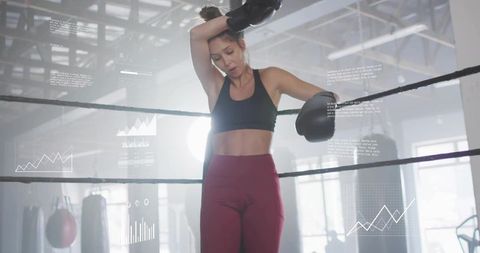 Female boxer leaning on ring ropes wiping brow after intense training with data overlays