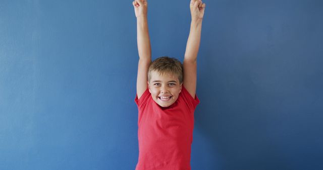 Young boy wearing a red shirt smiling with arms raised in front of a blue wall, expressing excitement and happiness. Great for illustrating themes of childhood joy, cheerfulness, and playful moments. Ideal for use in educational material, advertisements, family-oriented products, or any content related to kids and positivity.