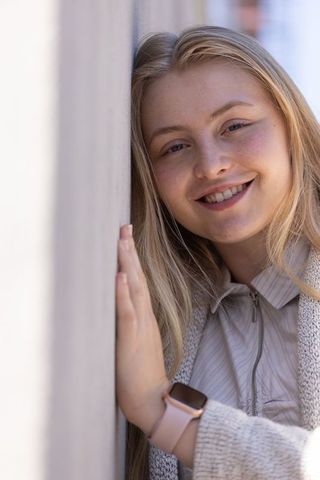 Young woman leaning on wall smiling while wearing knit cardigan and pink smartwatch