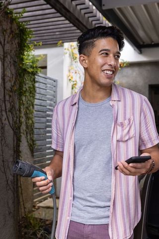 Young Man Charging Electric Vehicle and Using Smartphone in Carport