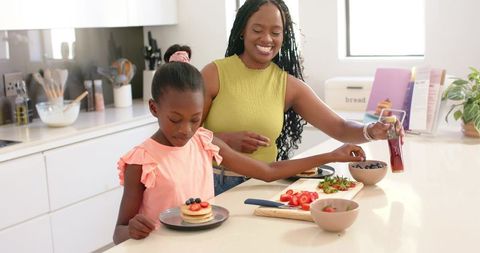 Smiling mother and daughter preparing pancakes on bright kitchen island, pouring syrup