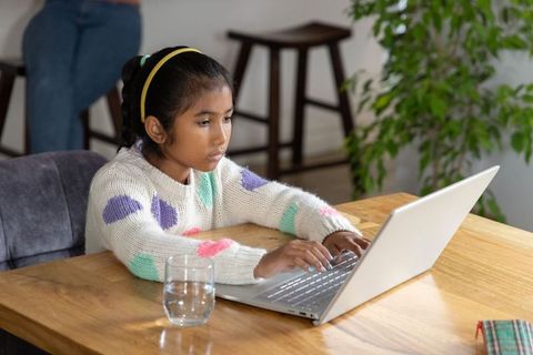 Asian girl studying on laptop at home desk wearing cozy sweater, remote learning