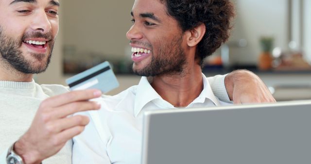 Two friends enjoying online shopping with a credit card and laptop in a domestic setting. Ideal for use in advertising for e-commerce, online banking services, credit card promotions, social media campaigns related to online shopping, or blogs about financial tips and friendship.