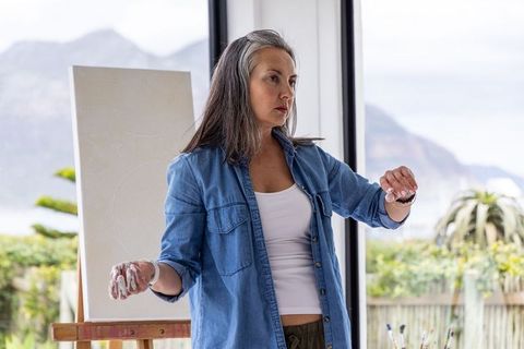 Senior woman artist assessing blank canvas in sunlit coastal studio with painted hands