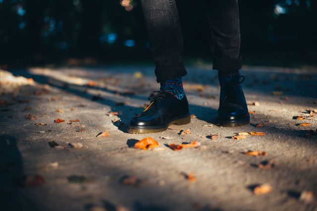 Showing lower legs of a person wearing black leather shoes and colorful socks standing on path covered with dry leaves in autumn park. This can be used for fashion, outdoor lifestyle, autumn themes, footwear advertisements, or seasonal promotions.