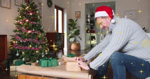 Middle-aged man wrapping christmas gifts at home wearing santa hat and cozy sweater