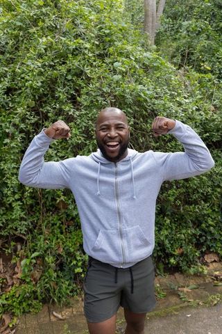 Smiling african american man flexing muscles on green trail wearing hoodie and shorts