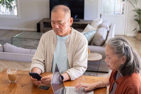 Senior asian couple using laptop and smartphone at sunlit wooden table for home banking
