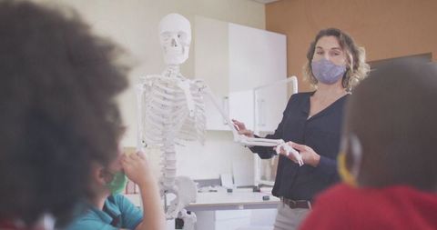 Teacher explaining human skeleton model to attentive schoolchildren wearing masks
